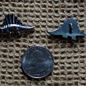 May include: Two silver-toned dinosaur buttons. One button is a side view of a dinosaur with spikes on its back. The other button is a top view of a dinosaur with spikes on its back. The buttons are on a brown textured surface.