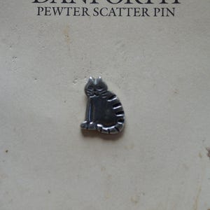 Könnte beinhalten: Eine silberne Zinn-Streupinne mit einer sitzenden Katze. Die Nadel befindet sich auf einer weißen Karte mit dem Text "Danforth Pewter Scatter Pin" und "Danforth Pewterers Ltd. Made in Middlebury, Vermont USA".