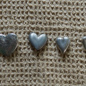 May include: Four silver heart-shaped charms of varying sizes. The largest charm has an intricate floral design.
