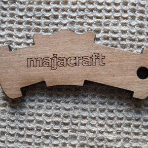May include: A light brown wooden tool with a curved, notched shape and a circular hole on the right side. The word majacraft is engraved in lowercase letters in the center of the object. The surface of the wood shows a natural grain pattern.