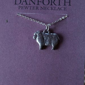 Könnte beinhalten: Eine silberne Zinnkette mit einem Schaf-Anhänger. Die Kette liegt auf einem violetten Hintergrund mit dem Text "DANFORTH PEWTER NECKLACE".