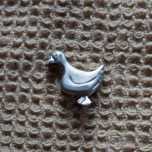 May include: A silver metal duck figurine with a detailed design. The duck is standing on a textured beige surface.