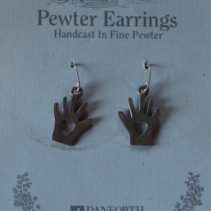 May include: A pair of pewter earrings shaped like hands with a heart cutout. The earrings are hand cast and have surgical steel posts and wires. The card reads "Pewter Earrings Handcast In Fine Pewter" and "Danforth Pewterers Maiden Vermont" with the copyright symbol and "©1990 Danforth Pewterers Ltd., Middlebury, VT Surgical Steel Posts And Wires".