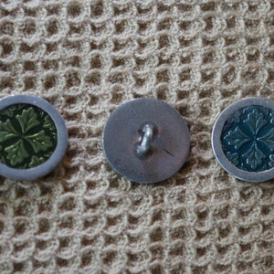 May include: Three round metal buttons with a silver finish. The buttons have a raised design of a four-leaf clover with a green or blue center.