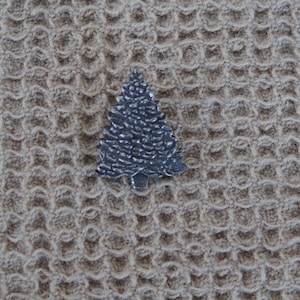 May include: A silver metal Christmas tree ornament in the shape of a pine tree. The ornament is on a beige textured background.