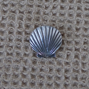 May include: A silver seashell charm with a detailed, ribbed texture. The charm is on a textured, beige background.
