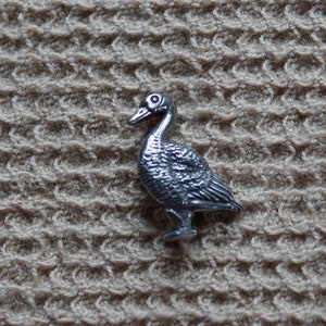 May include: A silver-colored metal goose figurine. The goose is standing on a textured beige surface.