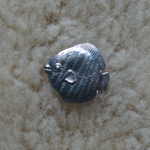 May include: A silver-colored fish charm with a detailed design. The charm is a stylized representation of a fish with fins and scales.