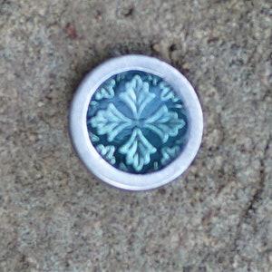 May include: A silver-colored round button with a blue glass center featuring a stylized floral design.