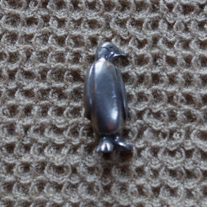 May include: A silver penguin figurine with a smooth, shiny finish. The penguin is standing upright with its wings tucked in and its head tilted slightly to the side.