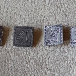 May include: Four silver-toned metal squares with a Greek key border. Each square features a different engraved image: a scarab beetle, a falcon, an owl, and a bee.