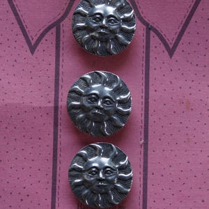 May include: Three pewter sun-faced button covers arranged vertically on a pink background. The buttons have a detailed sun design with a face in the center. The text "Button Covers" is at the top. The buttons are made by Danforth Pewterers.