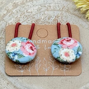 Könnte beinhalten: Zwei Haarspangen mit Blumenmuster und roten Gummibändern. Die stoffbezogenen Knöpfe haben einen hellblauen Hintergrund mit rosa und weißen Rosen und grünen Blättern. Sie werden auf einer braunen Karte mit dem Text "handmade with love" präsentiert.