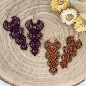 May include: Two pairs of macrame earrings are displayed. One pair is a deep burgundy, the other a warm orange-brown. Both feature a heart-shaped design with beaded accents and silver or gold-coloured hoops. The earrings are handmade.