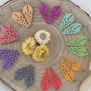 Può includere: Assortimento di orecchini in macramè fatti a mano in vari colori, tra cui rosa, viola, verde, arancione e grigio. Gli orecchini sono esposti su una superficie di legno con fiori secchi.