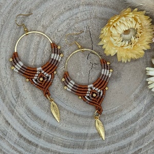 Op de afbeelding: Handgemaakte macramé oorbellen met goudkleurige ringen en bladbedels. De oorbellen hebben een geometrisch patroon in bruin, wit en beige, met kleine gouden kralen. Gepresenteerd op een houten oppervlak. Diameter ongeveer 6 cm.