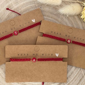 Könnte beinhalten: Rote Makramee-Armbänder mit Buchstabenperlen, präsentiert auf braunen Karten. Jedes Armband hat einen anderen Buchstaben: I, G und A. Auf den Karten steht "WEAR ME WITH" und ein kleines weißes Herz.