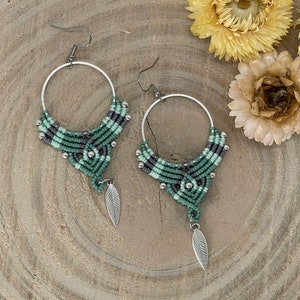 May include: A pair of green and gray macrame earrings with silver leaf charms. The earrings are hanging from silver hoops.