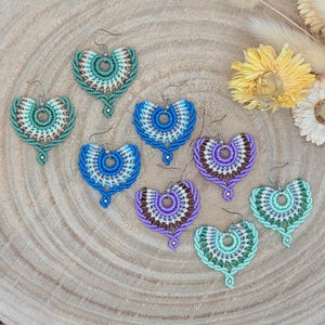 May include: A set of eight crocheted earrings in various colors, including green, blue, purple, and brown. The earrings are shaped like a fan and have a small circle in the center. They are displayed on a wooden surface.