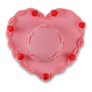 May include: A pink heart-shaped cake decorated with red candy hearts. The cake has a smaller pink cake on top with a bow.