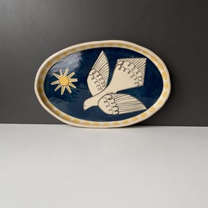 May include: An oval ceramic platter featuring a deep blue centre with a white bird illustration and a yellow sun. The rim is cream-coloured with a repeating pattern of yellow semi-circles. The surface has a glossy finish and irregular edges.
