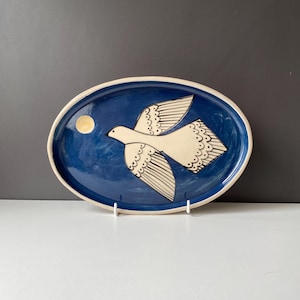 May include: An oval ceramic platter with a deep blue glazed surface featuring a hand-painted cream-coloured bird in flight. The bird has black line details on its wings and tail feathers. A small yellow circle representing the moon is visible.