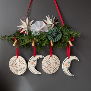 Pode incluir: Uma decoração de parede com um suporte de madeira adornado com ramos de pinheiro, estrelas de papel brancas e verdes e quatro ornamentos de cerâmica suspensos por fitas vermelhas. Os ornamentos retratam dois sóis e duas luas crescentes.