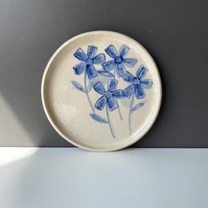May include: A white ceramic plate with a blue floral design. The plate features four stylized blue flowers with white centers and green stems.