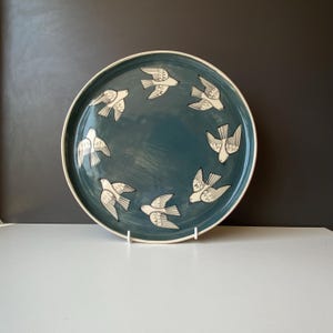 Op de afbeelding: Een ronde keramische bord met een diep blauwgroen glazuur, voorzien van een cirkelvormig patroon van zes vliegende witte vogels. De vogels zijn afgebeeld met eenvoudige zwarte lijndetails op hun vleugels en lichamen, gerangschikt in een cirkel.