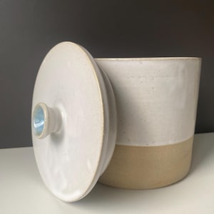 May include: A ceramic jar with a lid, in a neutral colour palette. The jar is cylindrical with a cream-coloured upper half and a tan lower half. The lid is off to the side, revealing the interior. The lid has a small, light blue handle.