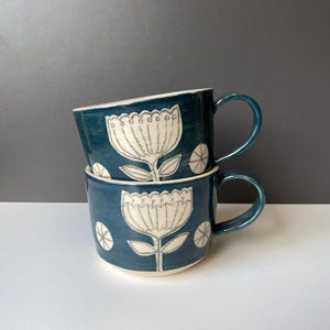 Op de afbeelding: Twee gestapelde keramische mokken in een diepe teal kleur. Elke mok heeft een wit bloemenontwerp met een geschulpte rand en kleine cirkelvormige accenten. De mokken hebben gebogen handvatten en een witte rand.