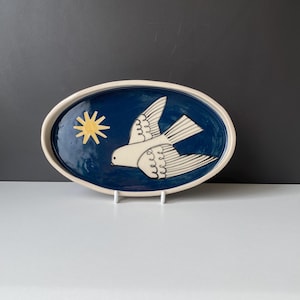 May include: An oval ceramic platter featuring a deep blue glazed interior with a hand-painted white bird in flight and a yellow star. The rim of the dish is a light cream colour, and the overall design is centred on the smooth, glossy surface.
