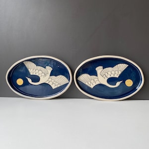 May include: Two oval ceramic platters featuring a deep blue glaze with a white crane illustration in flight and a yellow circular sun motif. Each platter has a raised white rim and a smooth, glossy finish, showcasing a matching artistic design.