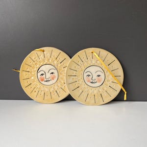 以下が含まれることがあります： 手描きの太陽の顔が描かれた2枚の円形セラミック製壁掛けプレート。ピンクの頬、黒い目、微笑む口が特徴です。クリーム色の表面には放射状の黒い線が施され、それぞれに吊り下げ用の黄色いリボンが付いています。