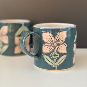 May include: Two ceramic mugs with a teal glaze and floral designs. The flowers are light pink with black outlines and green leaves. The mugs have a handle and a white interior.