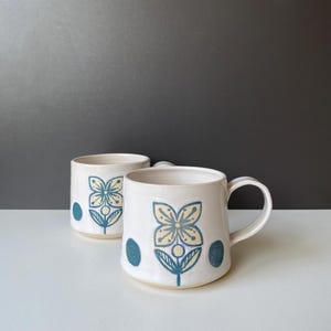 Op de afbeelding: Twee witte keramische mokken met een bloemmotief in blauw en geel. Elke mok heeft een grote bloem en twee blauwe stippen. De mokken hebben een handvat en een cilindrische vorm. De achtergrond is een verloop van wit en grijs.