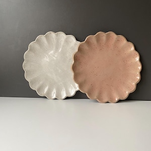 May include: Two ceramic plates with a scalloped design. One is off-white with speckled detailing, the other a muted pink with similar speckles. The plates are round and sit on a white surface against a grey backdrop.