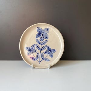 May include: A white ceramic plate with a blue floral design. The flowers are stylized and have four petals. The plate is round and has a slightly raised rim.