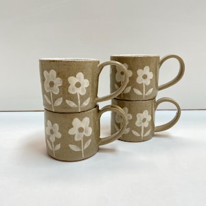 May include: Four ceramic mugs with a tan speckled finish and white floral designs. Each mug has a curved handle and a white interior. The mugs are stacked in two pairs, creating a visually appealing arrangement.