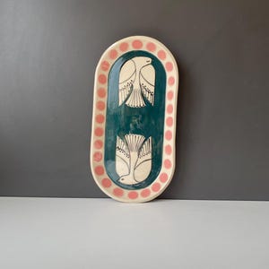 May include: An oval ceramic tray featuring a deep teal centre decorated with two white bird illustrations facing opposite directions. The rim is cream-coloured and accented with a repeating pattern of soft pink circular dots around the edge.