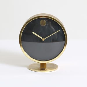 May include: A gold-toned desk clock with a black face and gold hands. The clock has a circular face and a round base. The clock's face is divided into two sections, with the top half being black and the bottom half being a darker shade of black.