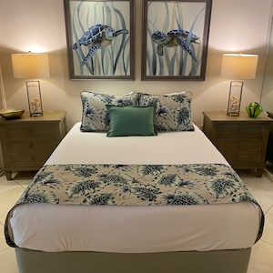 Könnte beinhalten: Ein Schlafzimmer mit einem Bett mit weißer Bettwäsche, ergänzt durch gemusterte Kissen und einen tropischen Blätterläufer. Über dem Bett hängen zwei gerahmte Gemälde von Schildkröten. Nachttische mit Lampen vervollständigen die Einrichtung des Raumes.