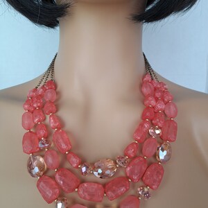 May include: A three-strand coral-colored beaded necklace. The necklace features large, faceted beads in varying shapes and sizes, interspersed with smaller, clear crystal beads. The necklace is displayed on a mannequin.