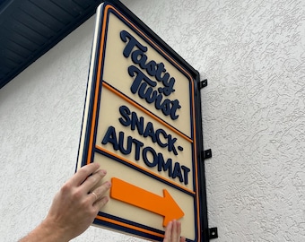 May include: Rectangular sign with the words "Tasty Twist SNACK-AUTOMAT" in dark blue lettering, with an orange arrow. The sign has a cream-colored background and is framed in black and orange. The sign is mounted on a textured white wall.