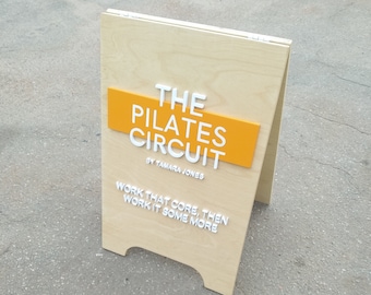 May include: A wooden A-frame sign with the text "THE PILATES CIRCUIT BY TAMARA JONES" in white letters on a yellow background. The sign also has the text "WORK THAT CORE, THEN WORK IT SOME MORE" in white letters on a wooden background.