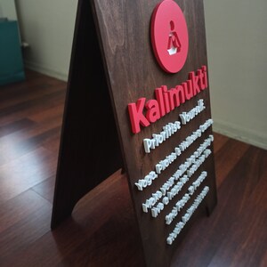 K&ouml;nnte beinhalten: Ein braunes, A-f&ouml;rmiges Schild aus Holz mit den Worten "Kalimukti" in roten Buchstaben und dem Text "Prioritise Yourself. Yoga, Pilates & Wellbeing. Make your wellbeing a priority. Live a happier, healthier life. One breath at a time. And find your inner peace." in wei&szlig;en Buchstaben.