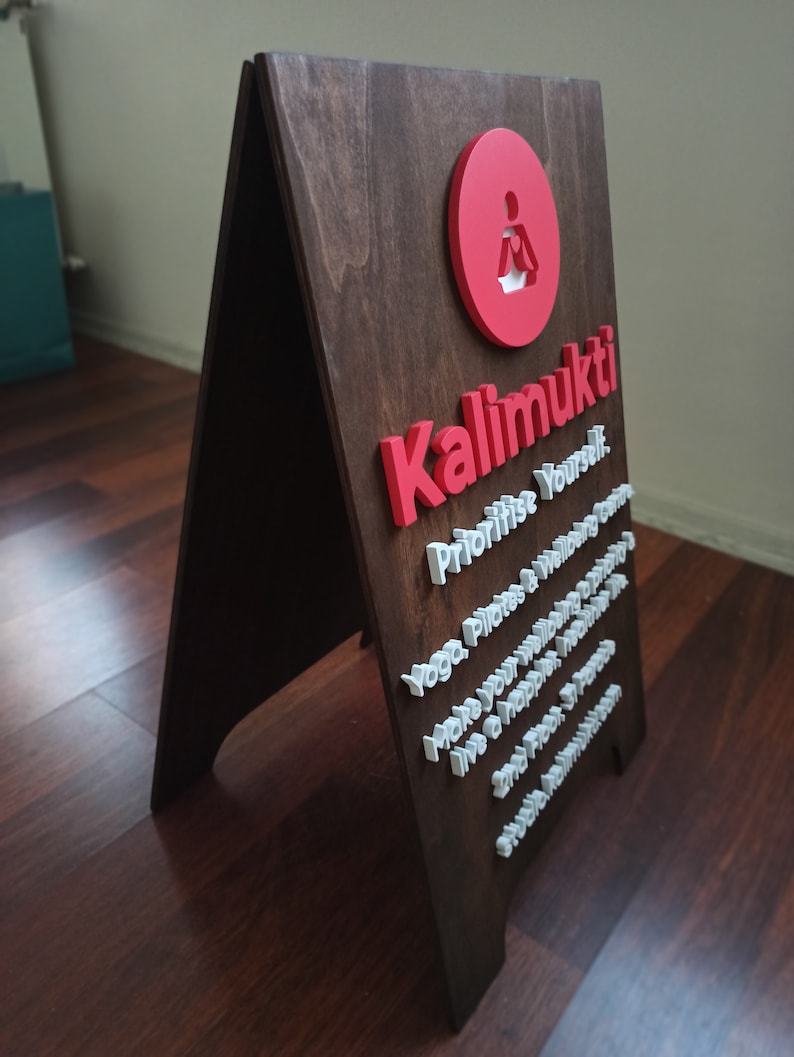 K&ouml;nnte beinhalten: Ein braunes, A-f&ouml;rmiges Schild aus Holz mit den Worten "Kalimukti" in roten Buchstaben und dem Text "Prioritise Yourself. Yoga, Pilates & Wellbeing. Make your wellbeing a priority. Live a happier, healthier life. One breath at a time. And find your inner peace." in wei&szlig;en Buchstaben.