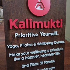 K&ouml;nnte beinhalten: Ein Schild aus Holz mit dem Text "Kalimukti" in roten Buchstaben. Das Schild enth&auml;lt auch den Text "Prioritise Yourself. Yoga, Pilates & Wellbeing Centre. Make your wellbeing a priority & live a happier, healthier life. 2nd Floor, 31 Parade studio.kalimukti.com" in wei&szlig;en Buchstaben.