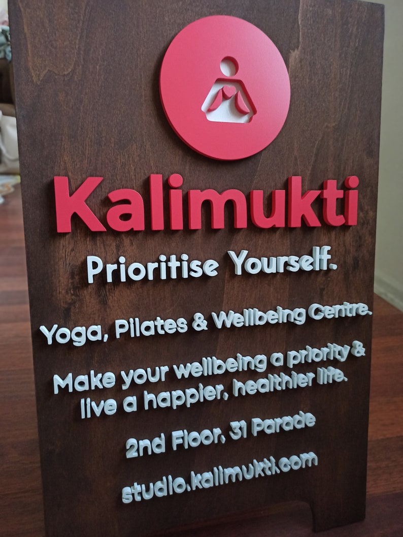 K&ouml;nnte beinhalten: Ein Schild aus Holz mit dem Text "Kalimukti" in roten Buchstaben. Das Schild enth&auml;lt auch den Text "Prioritise Yourself. Yoga, Pilates & Wellbeing Centre. Make your wellbeing a priority & live a happier, healthier life. 2nd Floor, 31 Parade studio.kalimukti.com" in wei&szlig;en Buchstaben.