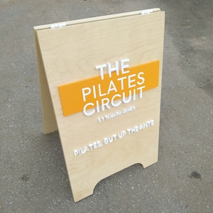 K&ouml;nnte beinhalten: Ein A-f&ouml;rmiges Schild aus Holz mit dem Text "THE PILATES CIRCUIT BY TAMARA JONES" in wei&szlig;en Buchstaben auf gelbem Hintergrund. Das Schild tr&auml;gt auch den Text "PILATES, BUT UP THE ANTE" in wei&szlig;en Buchstaben unten.